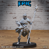 Skeleton king miniature wearing a jagged crown, raising a long twisted staff in one hand and clutching a skull-adorned open grimoire in the other, clad in armour and a flowing cloak. Sculpt by Epic Miniatures.