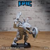 Frogfolk warrior lunging forward with a heavy hammer raised overhead in one hand and a round buckler held out in the other, wearing scale-textured armour over a padded jerkin with wrapped ankle bindings, wide jaw open in a full-toothed snarl, bony spines ringing the flat broad head. Sculpt by Epic Miniatures.