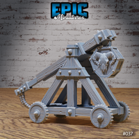 Orc war catapult with spiked wheels, wood-grain timber frame, iron rivets, coiled rope sling, and a carved ram-skull emblem on an octagonal shield plate. Sculpt by Epic Miniatures.
