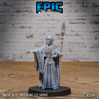 Robed elder male figure holding a tall beaded spear with diamond tip in one hand, opposite arm extended outward with fingers spread. Wears a fanned radial headdress, layered bead necklaces, and flowing ceremonial robes. Weathered, deeply lined face with a commanding expression. Sculpt by Epic Miniatures.