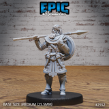 Bearded male warrior standing upright with a long spear rested across one shoulder, wearing layered scale armour, a decorative collar, pteryges at the waist, and lion-embossed greaves. Sculpt by Epic Miniatures.