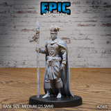 Muscular male tribal warrior standing upright, holding a tall feather-bound spear, wearing layered bead necklaces, a fringed wrap skirt, and a broad flowing cape. Feathered headdress atop rope-bound hair. Sculpt by Epic Miniatures.