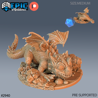 32mm scale resin miniature of a baby dragon hatchling by Epic Miniatures — unpainted digital render showing the creature in a low forward crouch on a rocky scatter base, wings half-raised, dorsal spines prominent, with a wide-eyed snarling expression and textured scales across the body; a blue d20 and top-down view inset provide scale reference