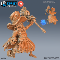Digital render of an unpainted resin voodoo goblin miniature at 32mm scale by Epic Miniatures, shown from the front and rear. She wears a wide-brimmed hat adorned with skulls, carries a tall staff topped with a feathered skull, holds a severed skull in her off hand, and has macabre trophies at her belt. A blue d20 die provides scale reference.