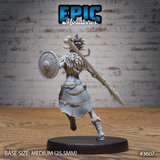 Rear view of the same unpainted grey resin tribal female warrior miniature, showing the detail of her feathered headdress, layered pelt cloak, and an ornate spear with a carved haft and beast-tooth tip, mounted on a medium round black base — by Epic Miniatures