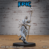 Rear three-quarter view of the unpainted grey resin Old Heretic miniature by Epic Miniatures, showing the flowing tattered robes, the back of the skull-topped staff, bundled spear hafts, and detailed wrap bindings on the forearms, on a medium 25.5mm round black base