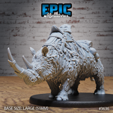 Front three-quarter view of an unpainted grey resin woolly rhino miniature on a 51mm oval base, showing shaggy fur texture, twin horns with metal cap on the primary horn, and detailed leather saddle with armour plates, bedroll, and pouches strapped across its back