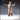 Digital render of Shopkeeper 01 miniature by Novaminis — a bald male figure in a long buttoned coat with rope belt and bow tie, arms thrown wide in an expressive merchant pose, viewed from the front