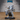 Unpainted grey resin 32mm miniature of a gnoll merchant in robes, crouching under a large pack stuffed with scrolls, tomes, and rope coils, holding cloth and rope in one clawed hand — front three-quarter view on a medium round base