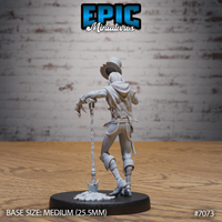 Grave digger miniature seen from behind, showing tattered layered vest, long hair beneath a battered top hat, and a ceremonial shovel with blade embedded in a skull-decorated earth mound base. Sculpt by Epic Miniatures.