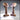 Digital render of two resin scatter terrain lecterns from Novaminis — one with an open book on the angled reading surface, one plain, both featuring riveted iron-banded bases and fluted wooden columns with rich grain texture, shown from the front at a slight angle
