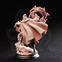 Photograph of the Aaron Drafthold Wandering Mage miniature from the rear, unpainted, showing the finely textured woven cloak billowing outward, runed shoulder clasp, and the swirling fire effect extending behind him over the rocky base