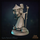 Agatha Saltwick miniature at a three-quarter rear angle: wicker basket detail visible, layered tattered robes, balance scale hanging from her outstretched right hand, clay pot and sack at her feet. Sculpt by Great Grimoire.