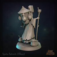 Agatha Saltwick miniature at a three-quarter rear angle: wicker basket detail visible, layered tattered robes, balance scale hanging from her outstretched right hand, clay pot and sack at her feet. Sculpt by Great Grimoire.