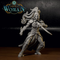 Painted grey resin miniature of a female dual-wielding rogue in a committed forward lunge, longsword levelled and short blade raised, ornate greaves and engraved bracers visible, windswept hair cresting at the crown. Sculpt by DnD Is A Woman.