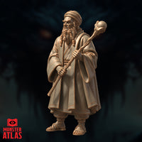 Elderly bearded male wizard in layered robes and turban, gripping a gnarled orb-topped staff in both hands, wearing sandals. Stern expression, braided beard. Sculpt by Monster Atlas.