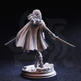 Rear view photograph of the printed Angela Cat-Eyes Hardvin miniature in unpainted grey resin, highlighting the detailed woven texture of her flowing cape, layered hair sculpt, and the ornate guard of one longsword extended behind her, mounted on a cobblestone rubble scenic base