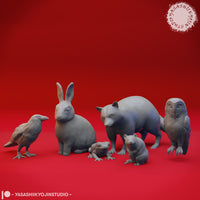 Digital render showing six of the woodland animal miniatures in closer detail — a raven, seated rabbit, foraging bear, frog, mouse, and barn owl — all unpainted grey sculpts against a red background by Yasashii Kyojin Studio