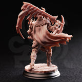 Rear view of the same unpainted resin Arcane Armour warrior miniature, showing the billowing tattered cape with detailed fabric folds, engraved back plate armour, and the twin serrated scythe-axe blades arcing above the figure against a dark background