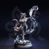 Unpainted grey resin miniature of Aria the Faithful tiefling sorceress, front three-quarter view — shows staff with dramatic translucent swirling energy effect, flowing cape, buckled belt with satchel, and rocky scenic base with pooling magical energy