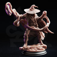 Front-facing view of Big Hat Harrison on a black display plinth, showing the full detail of his arcane medallion, belted coat, layered pouches, fleur-de-lis cape clasp, and the twin spiralling energy tendrils extending outward from both hands. Unpainted grey resin, 32mm scale.