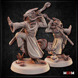 Two unpainted grey resin dragonborn monk miniatures shown side by side — the larger foreground figure strikes a raised-knee martial arts stance with staff swung overhead and one hand extended in a warding gesture, wearing layered flowing robes with a rope belt; the smaller background figure mirrors a similar pose from a different angle, both on detailed round bases with desert plants and broken stone