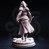 Rear view of the unpainted grey resin Delyna ro Unviel miniature on a round display base — revealing the textured chainmail hauberk, trailing torn skirts, rope-bound legs, spiked vambraces, devotional shield strapped to the belt, and the detailed sculpting on the rocky rubble base