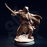 Elwahid Sword of the Sands miniature front view — unpainted resin desert elf in a deep hood, mid-lunge with curved blade extended, detailed carved tabard with sun emblem, on a round rocky base against a dark background.