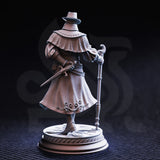 Rear three-quarter view of unpainted grey Ernest 'Flip-Flop' Dodd miniature showing the back of his quilted capelet, the flowing hem of his long embroidered coat with filigree detailing, and his right hand gripping a short cross-hilted blade. The decorative circular base is also clearly visible.
