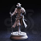 Front view of unpainted grey resin Ernest 'Flip-Flop' Dodd miniature on a decorative circular base. Clearly visible are his broad-brimmed hat, layered capelet with stud detailing, belted coat with scrollwork embroidery, ornate hooked staff, and tattooed forearm. Figure is mid-stride in a purposeful walking pose.