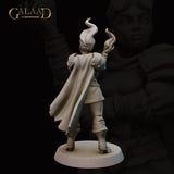 Unpainted grey resin miniature of a female human fire mage at 32mm scale, rear view. The back of her dramatic swept cape is visible in full, along with both arms raised with flame tendrils rising from her hands. Detailed pauldrons, fitted armour, and laced boots are clearly visible. Round base. Sculpted by Galaad Miniatures.