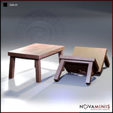 Two wooden table terrain pieces by Novaminis: one upright four-legged table with detailed wood grain, one dramatically overturned at an angle with chipped and splintered edge detail.