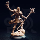 Studio photograph of the unpainted Hand of Seroca miniature from the rear, showing the back of her scaled hood and chainmail coif, layered back-plates, billowing skirt with woven texture detail, and the sun-star staff extending diagonally — the rocky terrain base clearly visible from this angle.