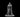 Grey resin digital render of a hooded human male warlock miniature at 32mm scale, standing upright in a long flowing cloak with mouth open in incantation, holding a skull focus outward in both hands, mounted on a plain round base