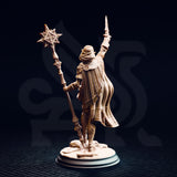 Unpainted resin miniature of Ilisyia the desert elf priestess, rear view. Shows the full-length flowing cape with panelled pleating, rear armour detailing including a circular embossed medallion at the shoulder, and the ornate shaft of her solar mace. Desert rubble base detail visible at foot level.