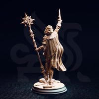Unpainted resin miniature of Ilisyia the desert elf priestess, rear view. Shows the full-length flowing cape with panelled pleating, rear armour detailing including a circular embossed medallion at the shoulder, and the ornate shaft of her solar mace. Desert rubble base detail visible at foot level.