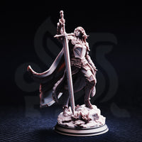 Front view of unpainted grey resin Jeanne du Lac miniature on a dark background — showing her full plate armour detail, raised greatsword, long flowing hair, and scenic rocky base with debris.