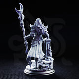 Kadna Glydrin by DM Stash — female elven warrior-mage standing on a vine-wrapped ruined stone base, holding a tall twisted staff topped with a crescent blade, wearing crescent pauldrons and filigree breastplate.