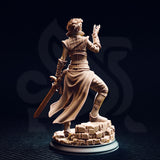 Rear view of the Karisia of Timberly half-elf Hexblade miniature — unpainted resin showing the woven texture of her long split tabard, laced bracers, elven longsword hilt detail, and the full sculpted rubble base, photographed against a dark studio background
