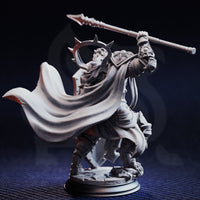 Rear three-quarter photograph of the unpainted grey resin Kigari Solheart miniature on a dark blue background, showcasing the dramatic flowing cloak caught mid-motion and the full sweep of the thorned halo crown. The reverse of the sculpted armour and the dynamic charging pose with raised spear are clearly visible, highlighting the quality of the sculpt's detail from all angles.
