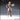 Front-facing digital render of Researcher 02 miniature by Novaminis — an unpainted resin sculpt of a confident woman in a button-up coat and knee-high boots, holding a large tome under her left arm with her right hand on her hip, braided hair swept back, 32mm scale