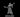 Unpainted grey resin miniature of a female lich in 32mm scale — a gaunt undead spellcaster with a spiked crown, tattered robes, one arm raised with clawed hand outstretched and the other holding a thick tome, mouth open mid-incantation, stood on a plain round base
