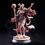 Female horned spellcaster miniature with curled ram-like horns and flowing tattered robes, holding an ornate staff topped with a spiky orb, posed on a bone-strewn base. Sculpt by DM Stash.