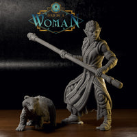 Photograph of a printed Xira Orc Monk miniature in unpainted grey resin alongside her bear companion, both placed on a wooden surface. The sculpt detail is visible across the robes, staff, braided hair, and bear fur texture.