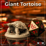 Photograph of a printed unpainted Giant Tortoise miniature in grey resin, shown on a tabletop gaming surface beside a large D20 dice for scale reference. The sculpt shows a detailed domed shell, wrinkled neck, and scaled limbs on a round scenic base.