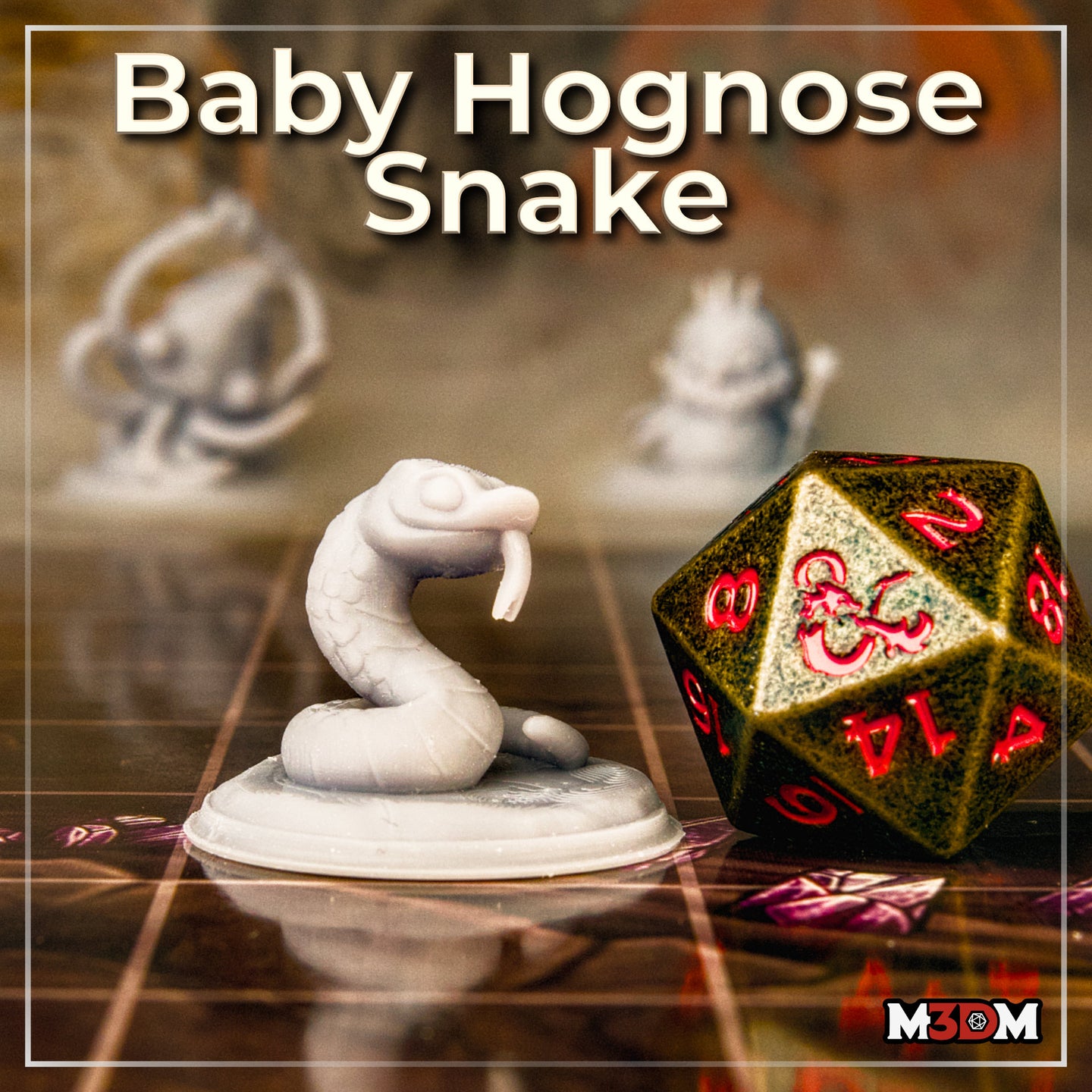 Photograph of a printed and unpainted Baby Hognose Snake miniature by Mia Kay M3DM on a gaming mat, shown in grey resin beside a large dark d20 die for scale — the coiled snake mini stands roughly one-quarter the height of the die, with a second blurred miniature visible in the background
