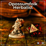 Photograph of the printed and unprimed Opossumfolk Herbalist miniature on a D&D battle mat alongside an oversized D20 die for scale — the 32mm grey resin mini shows strong detail in the joeys, scarf folds, and belt botanicals. M3DM branding visible in corner.