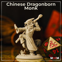 Single unpainted grey resin dragonborn monk miniature photographed beside a large D20 dice for scale — the figure balances on one leg with staff raised and free hand outstretched, showing draconic horns, trailing beard tendrils, and billowing robes with a textured desert base