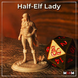 Photograph of a printed unpainted Half-Elf Lady resin miniature in grey, standing confidently with hand on hip, wearing a cloak and corset, on a scenic desert base with small succulents, shown beside a large D20 dice for scale — by M3DM