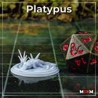 Unpainted grey resin platypus miniature printed at 32mm scale, shown on a gaming grid next to a large D&D d20 die for scale reference, with a blurred green foliage background